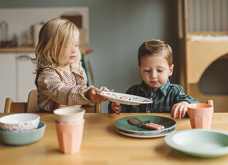 Twee kleine kinderen zitten aan een houten tafel met serviesgoed. Het blonde meisje reikt een leeg bord aan het jongetje, terwijl verschillende gekleurde kommen en bekers op de tafel staan in kinderopvang