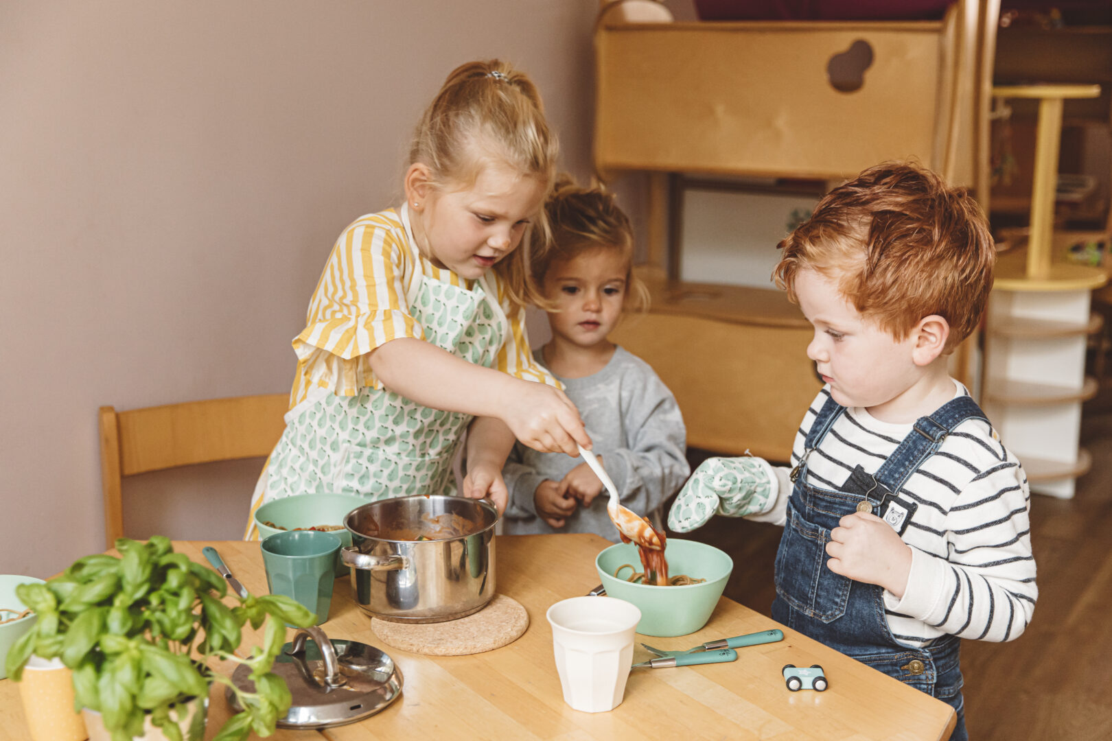 Drie jonge kinderen zijn samen aan het koken aan een houten tafel; het meisje in de gestreepte jurk serveert iets uit een pot terwijl het andere meisje en de jongen in de tuinbroek toekijken. Op tafel staan keukengerei en een plant.