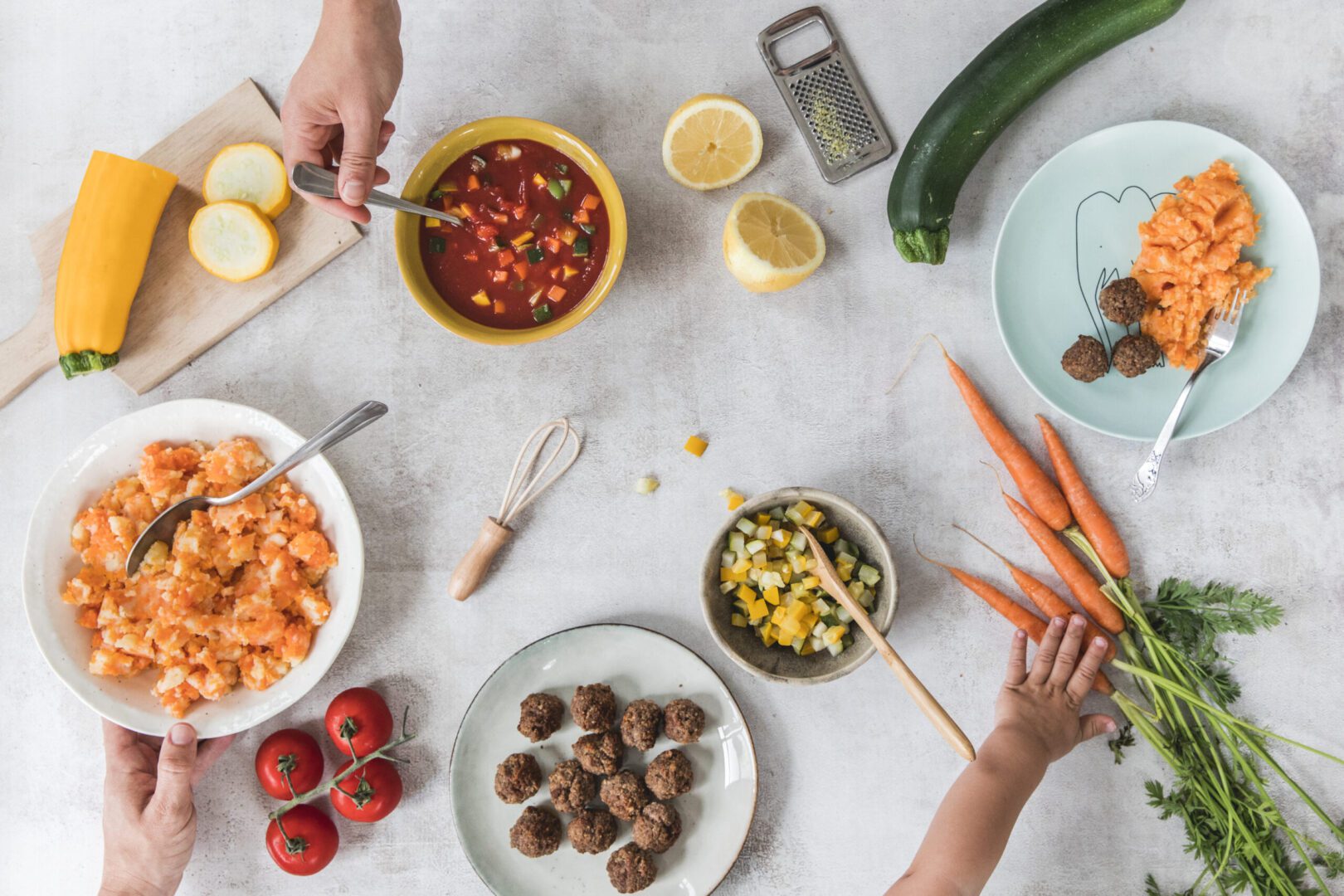 Een bovenaanzicht van een bezige keukentafel met een hand die een schaal met stamppot vasthoudt, een kom met verse salsa, gesneden courgette en een halve gele pompoen, een bord met wortelpuree en gehaktballetjes, en een kinderhand die naar wortels reikt