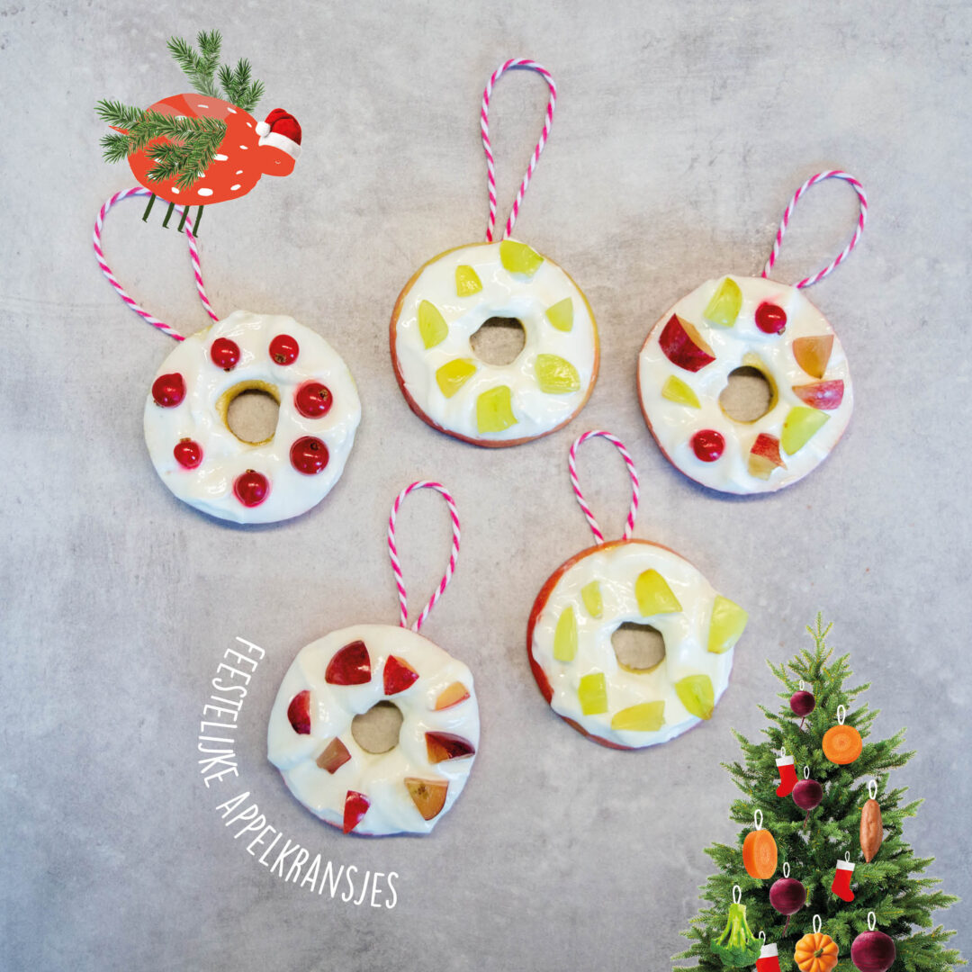 Feestelijke appelkransjes versierd met yoghurt en stukjes fruit als kerstversiering naast een kleine kerstboom gedecoreerd met fruit en groenten, met een illustratie van een vliegende kerstmuts boven een appel.