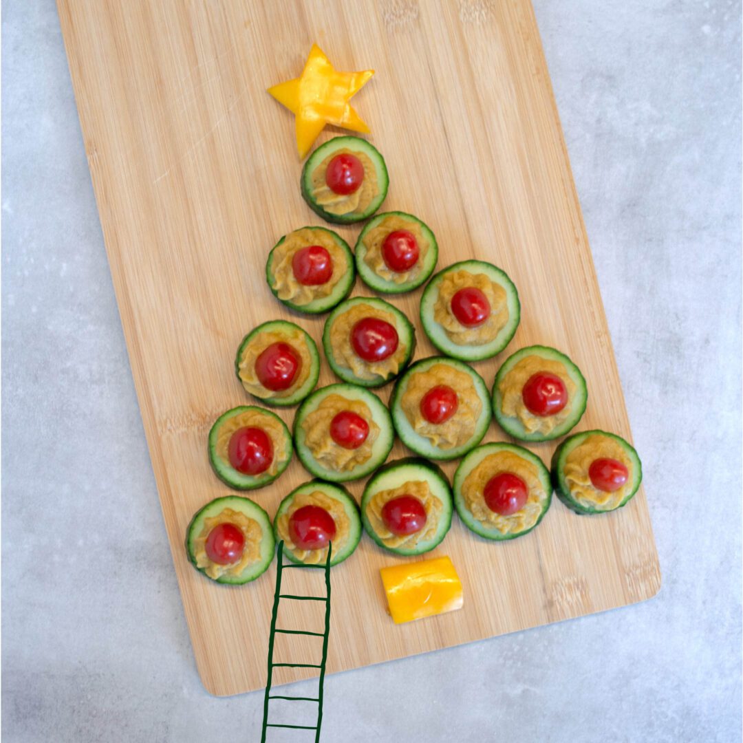 Een creatief kerstboomhapje gemaakt van plakjes komkommer en kerstomaatjes op pompoenspread, met een ster van kaas bovenop en een gele paprikavoet, gepresenteerd op een houten snijplank met een getekende ladder die naar de boom leidt.